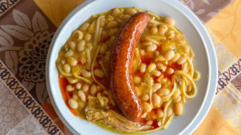 Porotos con riendas en un plato blanco, con una longaniza dorada encima